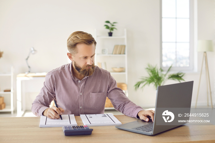 Serious businessman calculates company’s internal accounts using laptop and calculator. Focused man checks financial documents and plans budget while sitting at his workplace in office or at home.