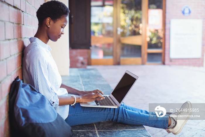 Attentive schoolgirl sitting against brick wall and using laptop