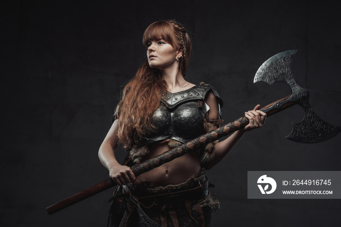 Armed with huge axe and armoured woman viking with long brown hairs poses in dark background.