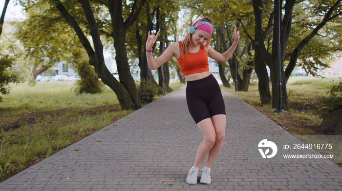 Athletic fitness sport runner girl training exercise, listening music on headphones dancing to camera having fun. Workout cardio in park. Young woman jogger enjoying funny dance. Active sportswoman