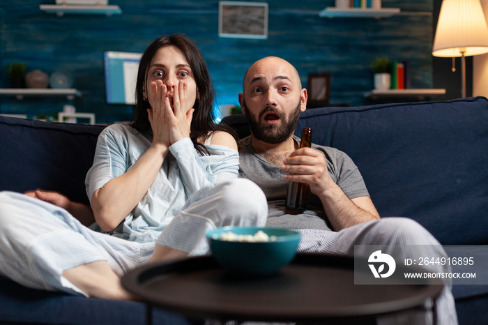 Confused shocked couple watching TV movie at night and eating popcorn, drinking beer having fear facial expression. Focused astonished people sitting on confortable couch chilling together