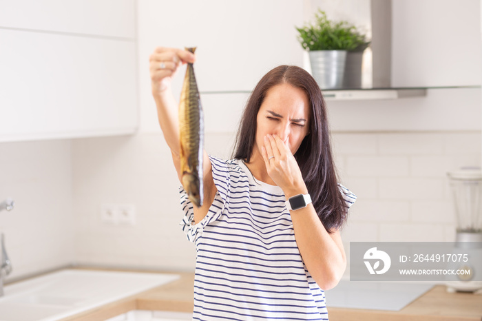 Woman disgusted from a smell of a fish she holds in hand holds her nose standing in the kitchen at home