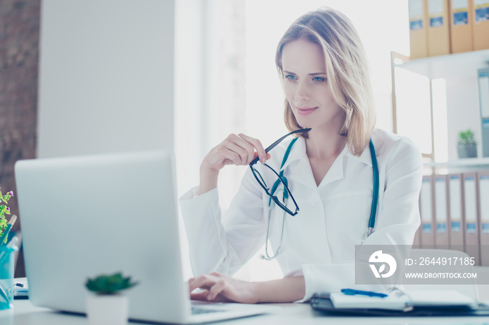 Concept of using modern technology at hospitals. Portrait of smiling charming female doc with glasses in hands, she is using her laptop for writing prescriptions