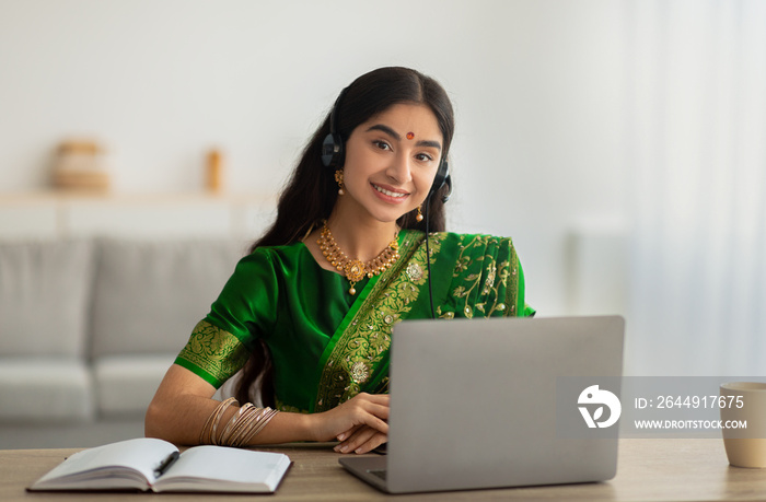 Indian woman in sari dress wearing headphones, having online meeting or web conference on laptop at home