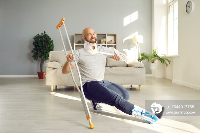 Happy man with multiple physical injuries having fun while recovering at home. Crazy, funny male patient with broken neck, sprained foot and injured arm moving and dancing with crutch in living room