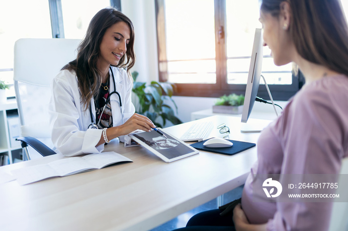 Pretty young woman gynecologist using digital tablet for showing the ultrasound to her pregnant patient in the clinic.