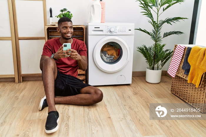 Young african american man using smartphone waiting for washing machine looking away to side with smile on face, natural expression. laughing confident.