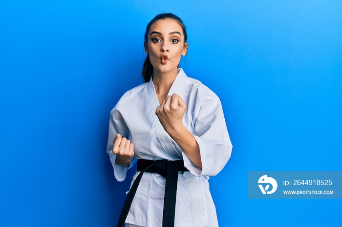 Beautiful brunette young woman wearing karate fighter uniform with black belt doing attack pose making fish face with mouth and squinting eyes, crazy and comical.