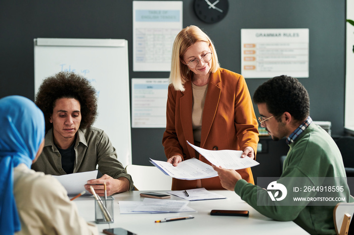 Happy mature blond teacher passing papers with grammar tests to young multicultural students sitting around table in classroom