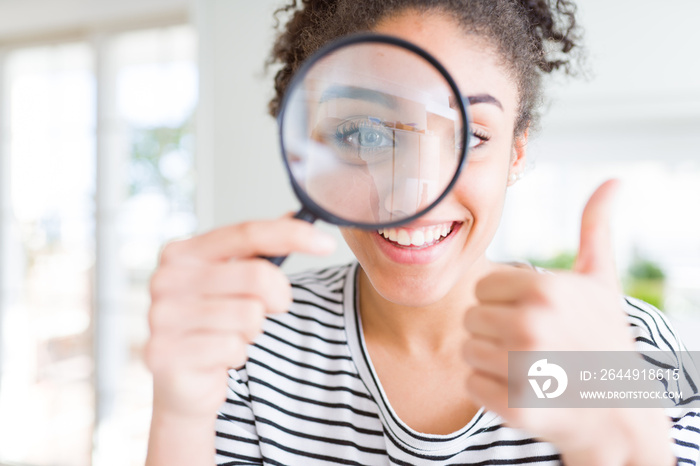 Young african american woman looking through magnifying glass happy with big smile doing ok sign, thumb up with fingers, excellent sign