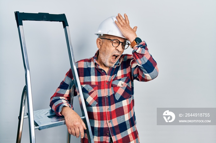 Handsome mature handyman close to construction stairs wearing hardhat surprised with hand on head for mistake, remember error. forgot, bad memory concept.