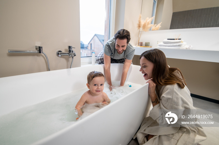 Child looking at camera in bathtub and parents watching