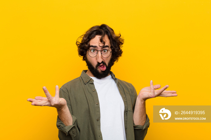 young crazy man shrugging with a dumb, crazy, confused, puzzled expression, feeling annoyed and clueless against yellow wall