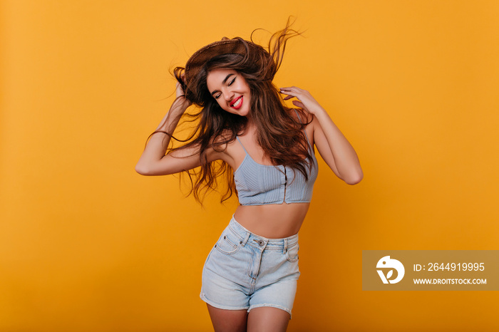 Ecstatic girl with inspired face expression dancing on yellow background. Studio shot of glamorous tanned female model in trendy shorts enjoying photoshoot.