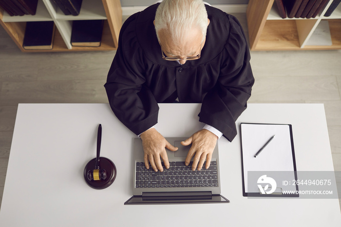 Law consultation and legal advice online: Professional senior judge, attorney or lawyer sitting at desk with laptop, doing web research or answering client’s question on website. High angle from above