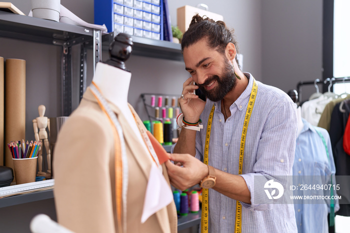 Young hispanic man tailor talking on smartphone standing by manikin at atelier