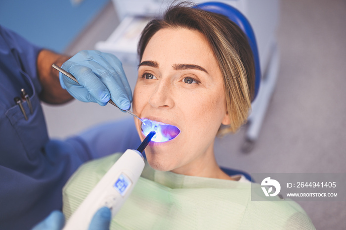 Dentist doctor using dental curing light equipment for filling, after examining a patient’s teeth in dentistry office. Stomatology and health care concept. Close up young happy woman in dental chair