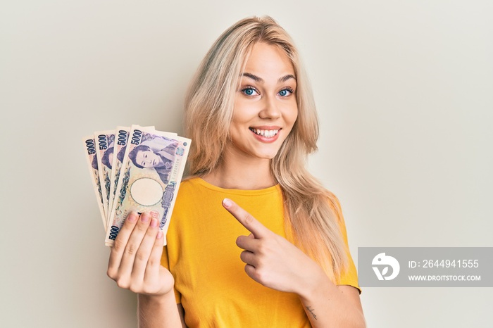 Beautiful caucasian blonde girl holding 5000 japanese yen banknotes smiling happy pointing with hand and finger