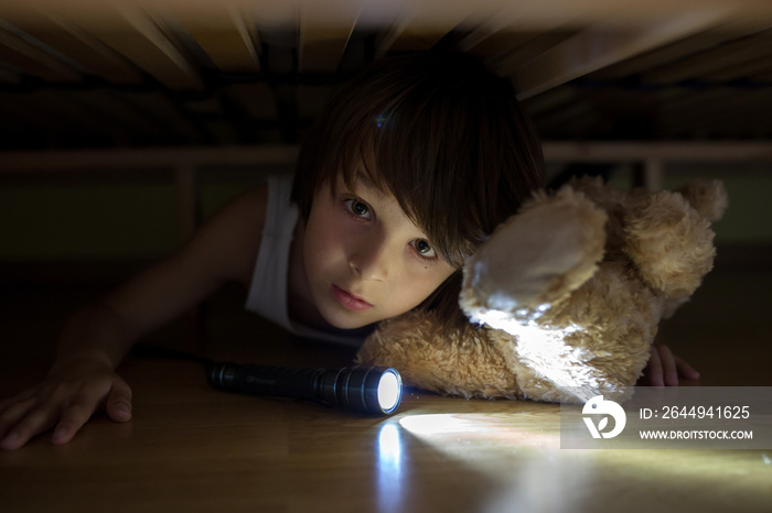 Little child, hiding under the bed, hugging teddy bear and holding flashlight,