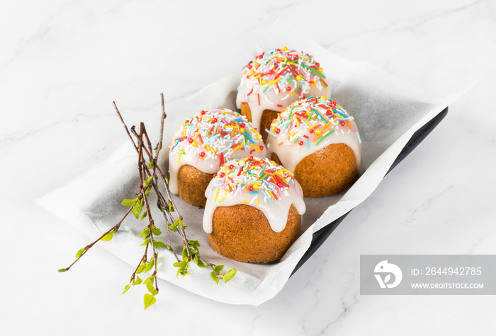 Easter small cakes with lemon sugar glaze, decorated with multicolored confectionery sprinkles, with spring birch twigs, on a serving plate. White background