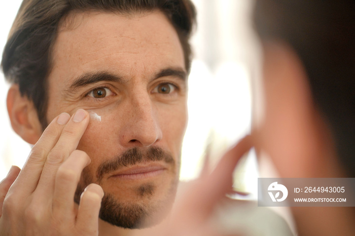 Handsome man applying face cream looking in mirror