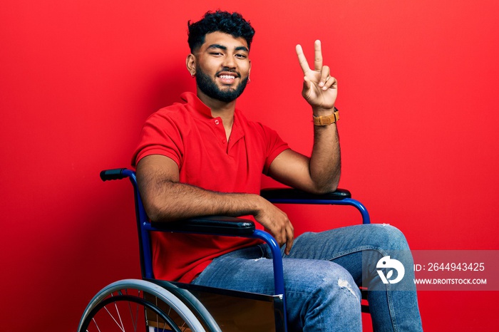 Arab man with beard sitting on wheelchair smiling looking to the camera showing fingers doing victory sign. number two.