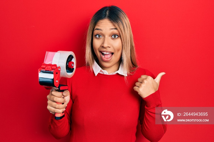 Beautiful hispanic woman holding packing tape pointing thumb up to the side smiling happy with open mouth