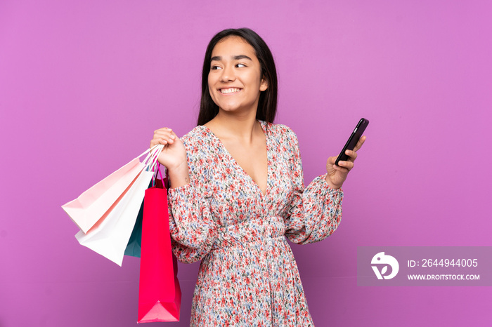 Young Indian woman isolated on purple background holding shopping bags and a mobile phone