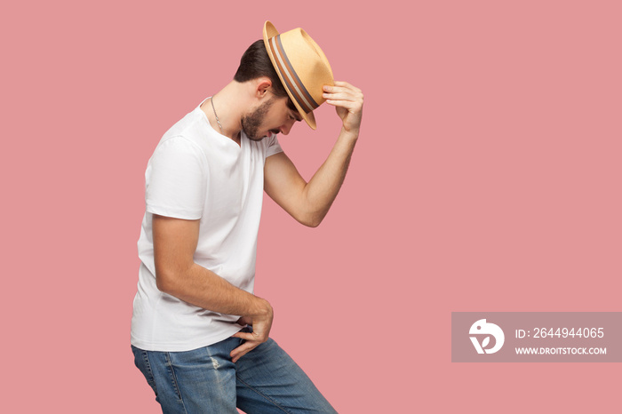 Profile side view portrait of bearded young dancer man in white shirt holding his hat and standing in michael jackson dancing pose. indoor studio shot, isolated on pink background copyspace.