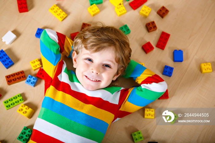 Little blond child playing with lots of colorful plastic blocks indoor. Kid boy wearing colorful shirt and having fun with building and creating