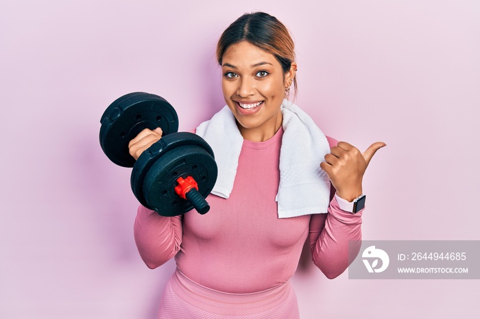 Beautiful hispanic woman wearing sportswear using dumbbells pointing thumb up to the side smiling happy with open mouth