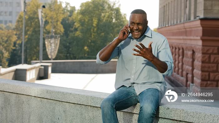 Worried nervous African American man in shirt and jeans holding mobile phone. Stressed frustrated client of retirement age talking to service sitting outdoors Customer dissatisfied with poor serve