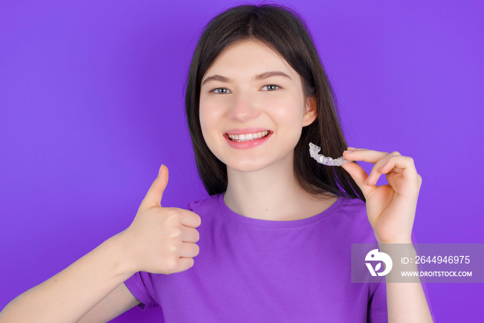 young beautiful Caucasian girl wearing purple T-shirt over purple background holding an invisible braces aligner and rising thumb up, recommending this new treatment. Dental healthcare concept.