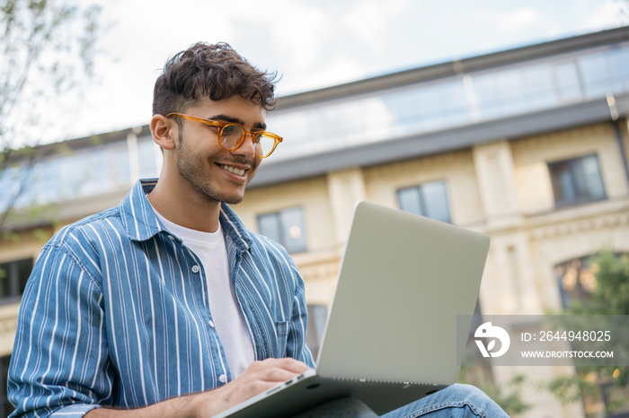 Portrait of happy Indian programmer using laptop computer, internet, working freelance project online, sitting in park. Asian student studying, learning language, using modern technologies outdoors