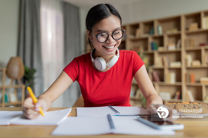 Glad teenage chinese girl student in glasses study at home, typing on phone in living room interior