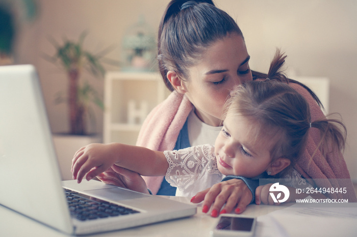 Young mother at home kissing her daughter and using laptop together.