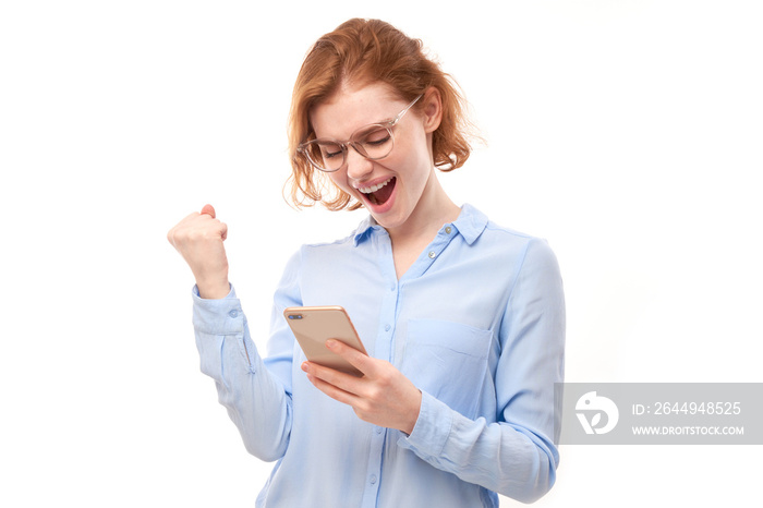 lucky smiling redhead girl holding smartphone in business shirt with glasses isolated in studio received joyful message, celebrating victory