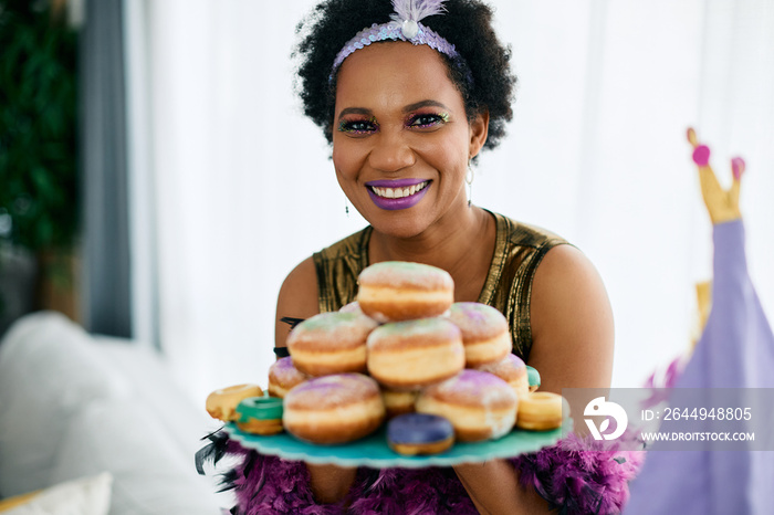 Happy African American woman wears Mardi Gras costume and make-up while holding plate of donuts at home.