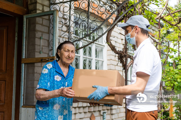 Young male volunteer in mask gives an elderly woman boxes with food near her house. The son helps a single elderly mother. Family support, caring. Quarantined, isolated. Coronavirus covid-19