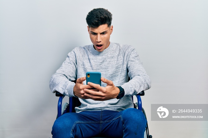 Young hispanic man sitting on wheelchair using smartphone afraid and shocked with surprise and amazed expression, fear and excited face.