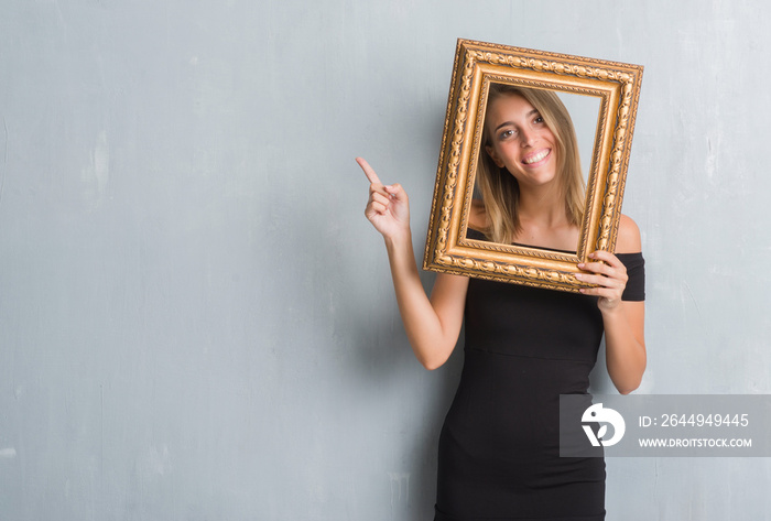 Beautiful young woman over grunge grey wall holding vintage frame very happy pointing with hand and finger to the side