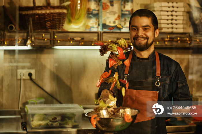 Smiling chef in jeans apron jumbling up vegetable salad in metal bowl. Salad consisting of tomatoes, cucumbers, paprika. Background of professional restaurant kitchen with special kitchenware.