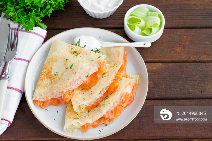 Thin pancakes or crepes with smoked salmon, cream cheese, cucumber and dill on a dark wooden background. Selective focus.