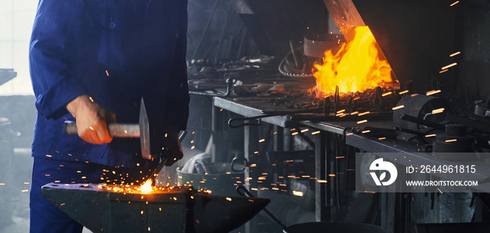 Close up of experienced blacksmith forging metal with heavy hammer on anvil. Man in blue overalls working with steel heated to high temperature.