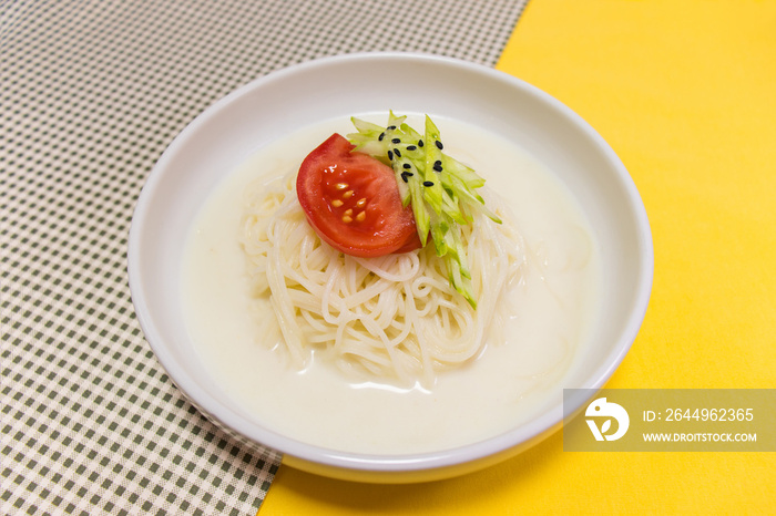 Korean summer food  Kongguksu  : Noodles in cold soybean soup, Cold bean soup noodles topped with sliced tomato and shredded cucumber in white bowl on green checkered and yellow tablecloth background.