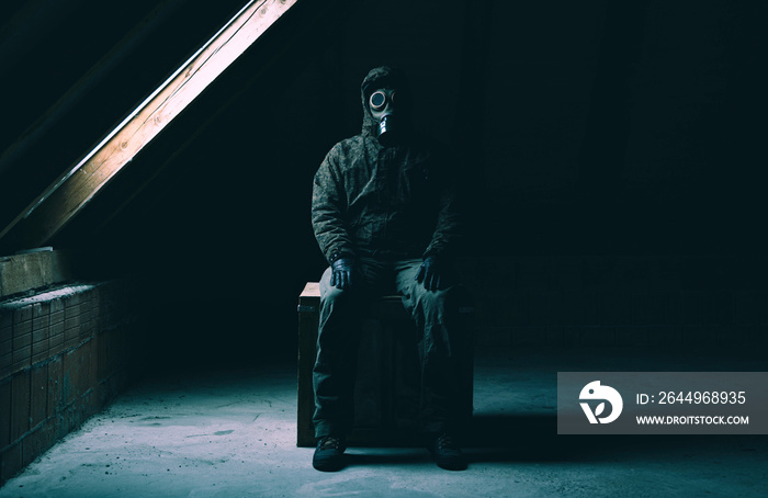 A man in gas mask and military-style clothes sitting on a wood box next to an attic window and looking into the camera.