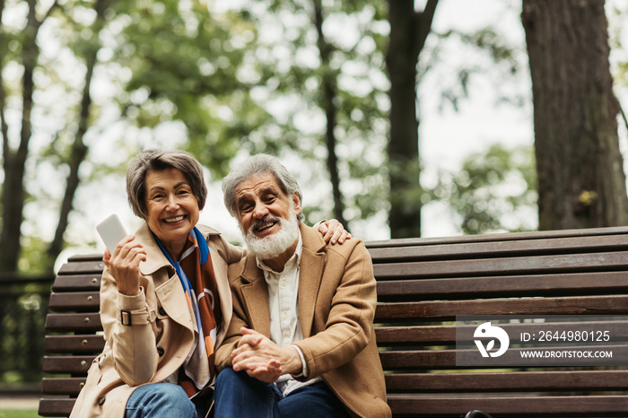 happy senior woman smiling while holding smartphone and sitting with bearded husband in coat on bench.