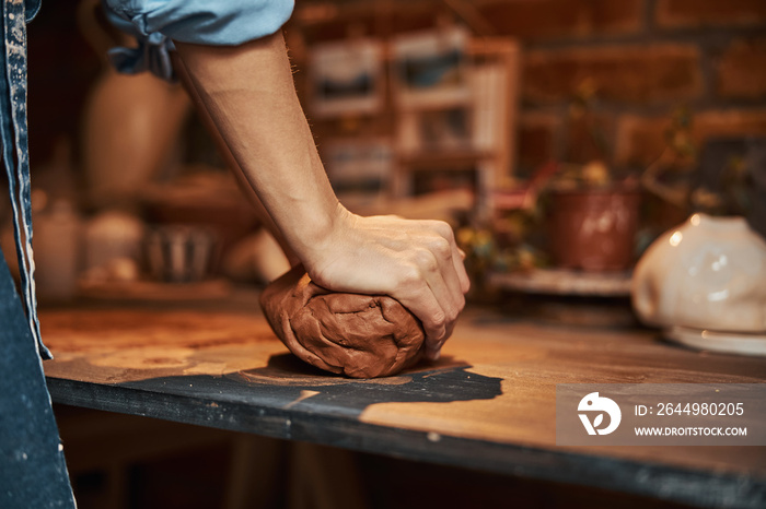 Unrecognized woman sculptor in apron standing near the work desk while shaping fresh clay in pottery studio