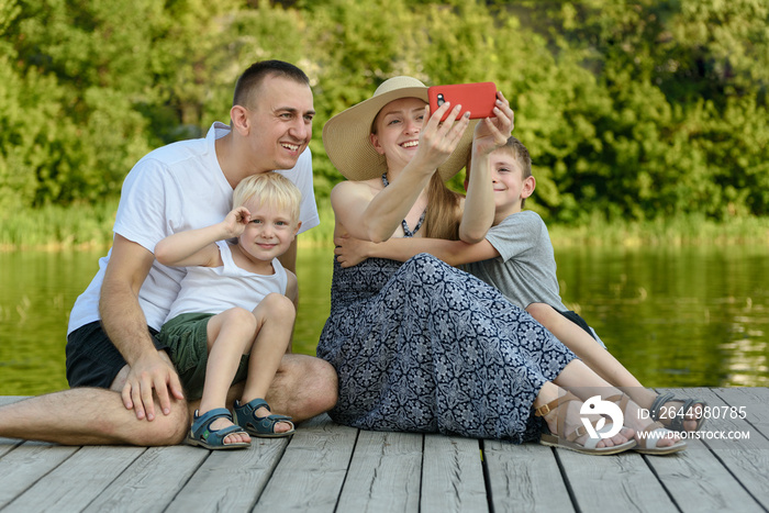 Happy young family, father mother and two little sons are sitting and taking selfies on the river pier