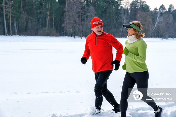 Smiling senior couple jogging in snowy winter park. Elderly wife and husband doing healthy exercise outdoors. Active lifestyle concept.